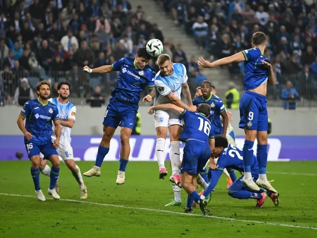 Câțiva jucători de fotbal în echipamente albastre și albe concurează pentru un balon în aer pe un teren de fotbal verde.