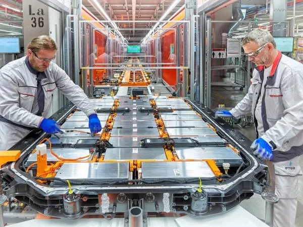 Doi bărbați cu mănuși albastre lucrează la o platformă metalică plată care conține componente electrice și cabluri portocalii într-un mediu industrial.