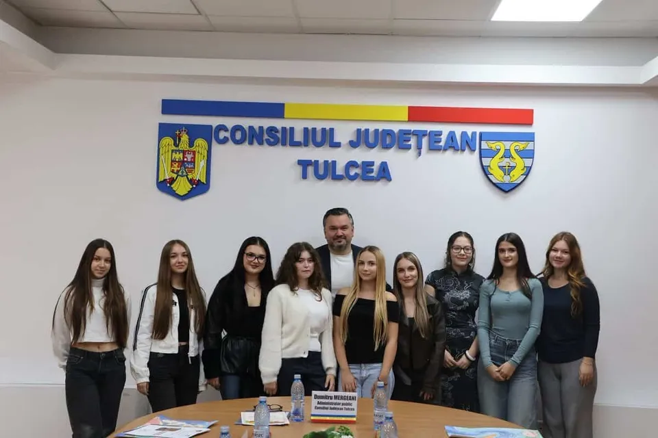 Un grup de nouă persoane stă în picioare în fața unui panou alb cu inscripții albastre și două embleme colorate.