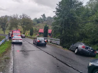 Un autoturism negru este oprit pe un drum asfaltat umed lângă o mașină de poliție albă și o ambulanță roșie sub un cer înnorat.