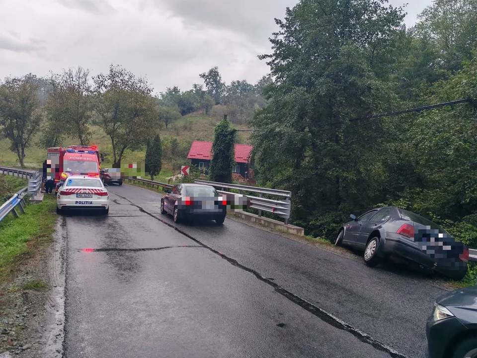 Un autoturism negru este oprit pe un drum asfaltat umed lângă o mașină de poliție albă și o ambulanță roșie sub un cer înnorat.
