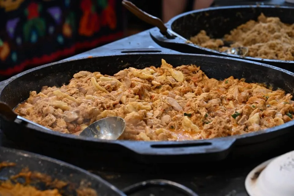 Două tăvi mari de fier conțin preparate culinare găsite pe o suprafață plană, fiind vizibile o lingură și un tacâm metalic.