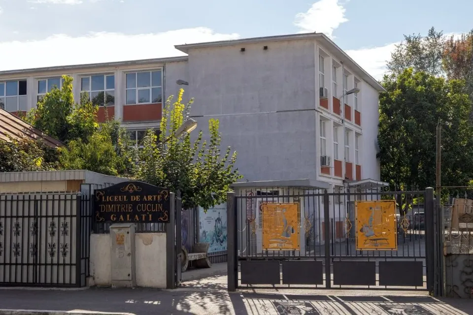 O clădire cu ferestre mari și panouri portocalii este situată în spatele unui gard metalic negru și a unui zid de beton.