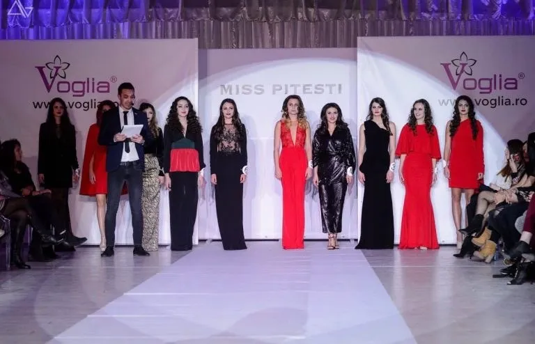 O serie de femei în diverse ținute de seară stau pe un podium alb, lângă un bărbat cu o foaie în mână.