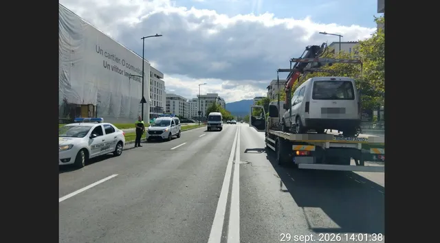 Un autovehicul alb este ridicat de pe asfalt de către un camion cu platformă pe un drum urban cu marcaje albe.