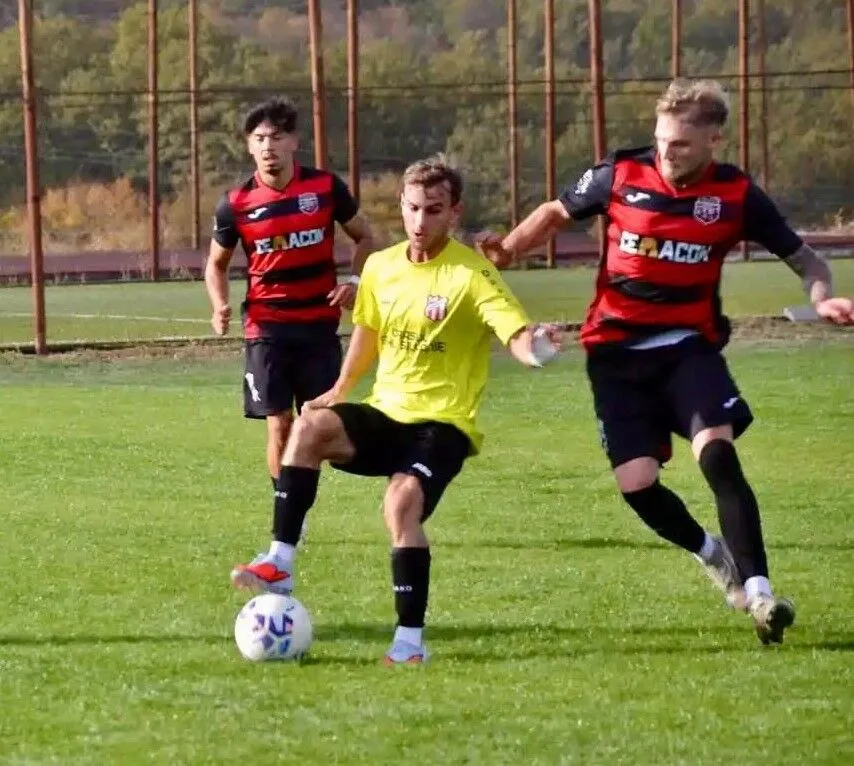 Un jucător în echipament galben conduce mingea pe un teren de fotbal, fiind urmărit de doi adversari în echipamente roșii și negre.