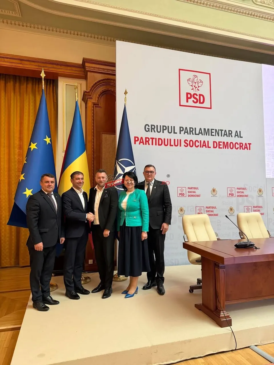 Cinci persoane stau în picioare în fața unui panou alb cu logoul PSD și steaguri, lângă o masă de lemn cu scaune albe.