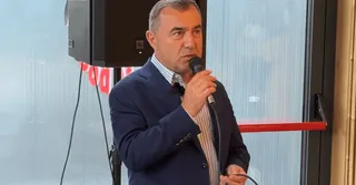 Un bărbat cu părul scurt și sacou albastru ține un microfon negru în mână în fața unui difuzor și a unei ferestre.