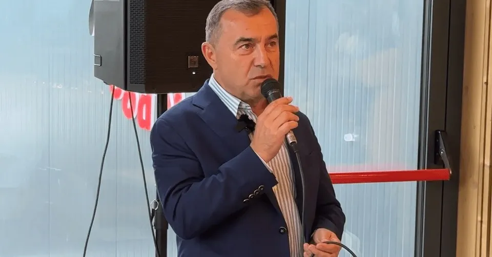 Un bărbat cu părul scurt și sacou albastru ține un microfon negru în mână în fața unui difuzor și a unei ferestre.