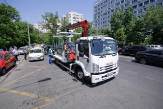 Un camion alb cu braț hidraulic transportă un vehicul pe o platformă într-o stradă cu mașini parcate și clădiri înalte în fundal.
