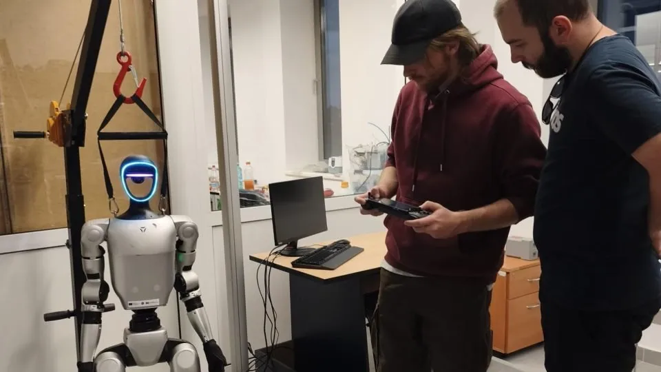 Un robot umanoid alb și gri stă suspendat de un cârlig, în timp ce doi bărbați stau lângă un birou cu monitor și tastatură.