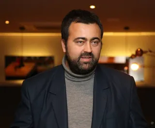 Un bărbat cu barbă și păr scurt poartă un sacou închis la culoare peste un pulover cu guler înalt gri.