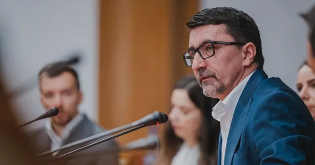 Un bărbat cu ochelari și sacou albastru stă în prim-plan, în timp ce alte persoane apar neclare în fundal lângă microfoane.