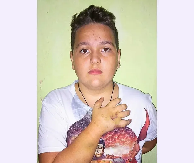 Un adolescent cu părul scurt și tricou alb cu imprimeu portând un lăn de sub gât își ține mâna pe piept.