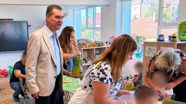 Un bărbat într-un sacou deschis stă în picioare lângă un grup de femei și copii într-o încăpere luminoasă cu rafturi de jucării și ferestre mari.