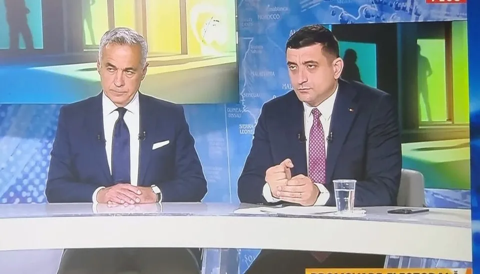 Doi bărbați în costume scurte stau la o masă albă în fața unui fundal de studio televizat.