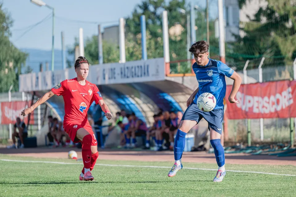 Un jucător în echipament albastru controlează un fotbal, în timp ce un alt jucător în echipament roșu se află în apropiere pe un teren de fotbal.