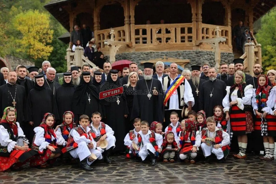 Un grup de oameni, inclusiv clérul în haine negre și copii în costume populare, pozează în fața unei construcții din lemn.