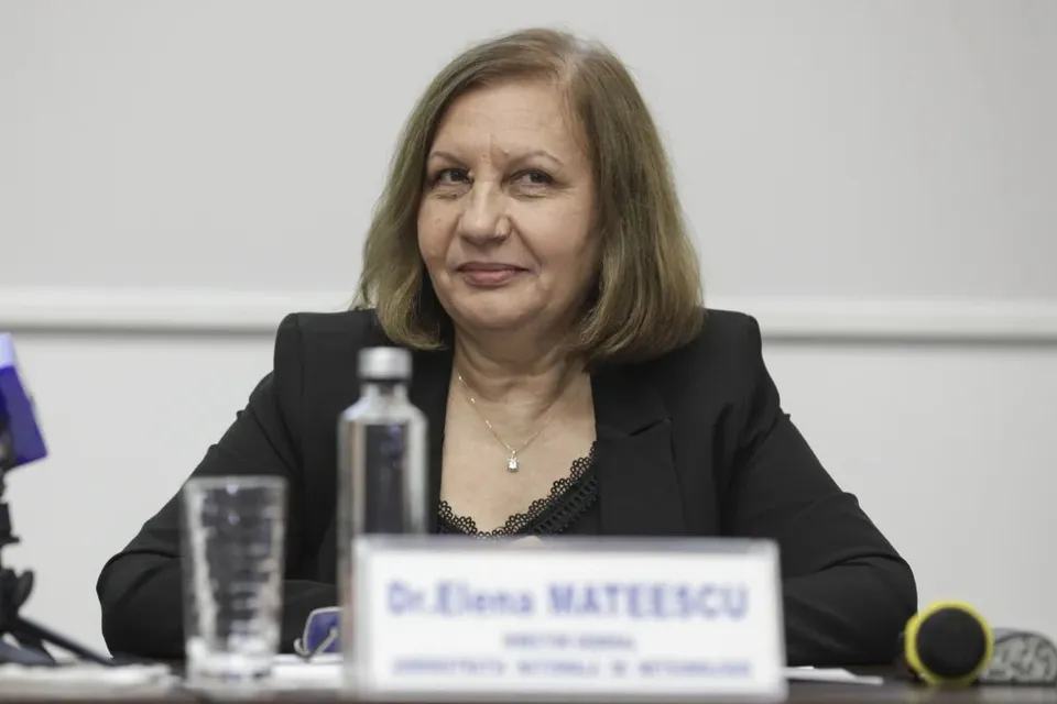 O femeie cu părul scurt și blond, purtând o jachetă neagră și un colier, stă la o masă lângă o sticlă și un pahar.