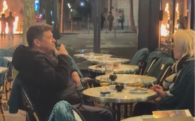 Un bărbat și o femeie stau la mese rotunde de cafea într-un local, în timp ce flăcări ard în fundalul străzii de afară.