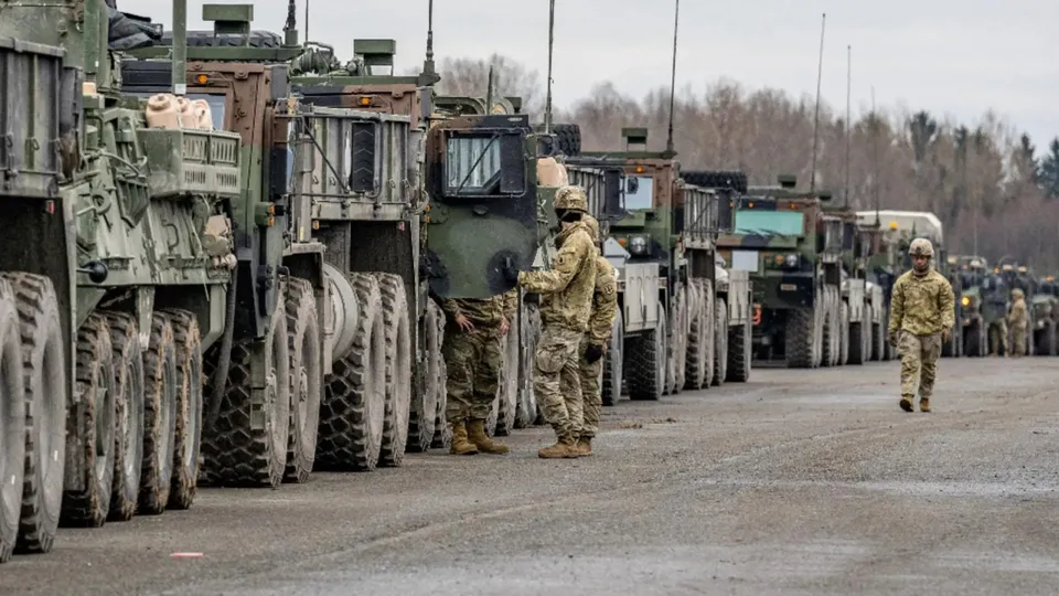 O linie de vehicule militare verzi este parcată pe un drum, în timp ce câțiva militari în uniforme de camuflaj stau lângă acestea.