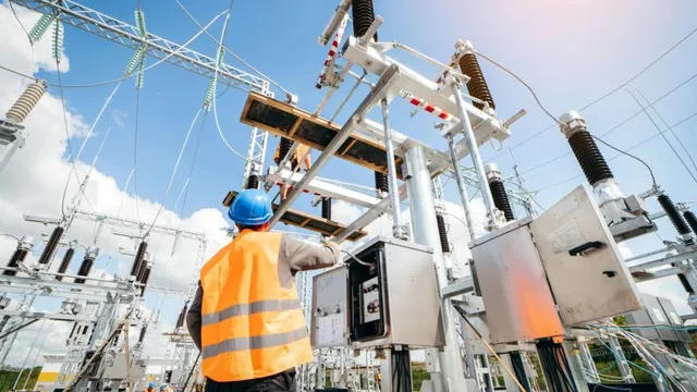 Un lucrător cu cască albastră și vestă reflectorizantă portocalie lucrează la echipamente electrice de înaltă tensiune sub un cer înnorat.