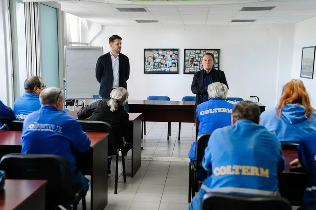 Un grup de oameni stă la o masă într-o încăpere cu pereți albi, în timp ce doi bărbați stau în picioare în fața lor.