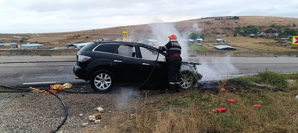 Un autoturism negru stă pe marginea unui drum lângă un pompier care poartă cască și echipament de protecție, în timp ce fumul se ridică din vehicul.