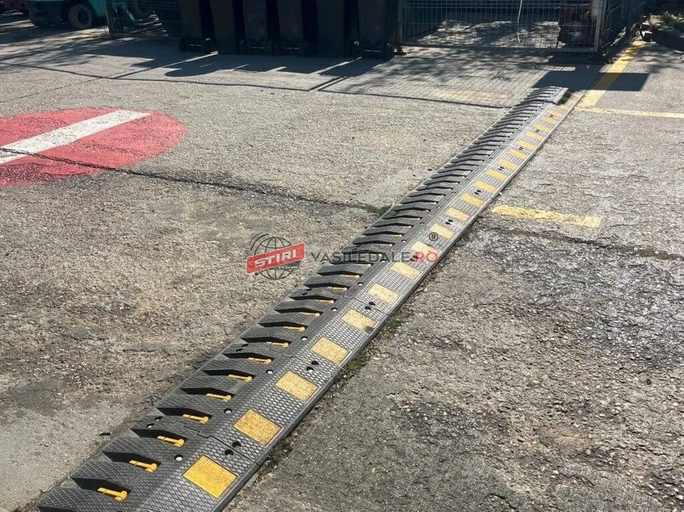 Un dispozitiv de tip bumper din cauciuc neagră cu marcaje galbene este montat pe o suprafață asfaltată lângă o zonă marcată cu vopsea roșie.