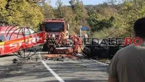 Un camion roșu și un autoturism negru sunt situate pe un drum asfaltat lângă vegetație abundentă.
