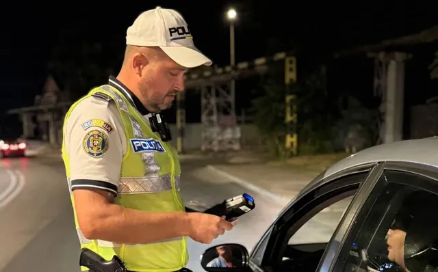 Un agent de poliție în uniformă și vestă reflectorizantă folosește un dispozitiv electronic lângă fereastra deschisă a unei mașini de culoare argintie noaptea.