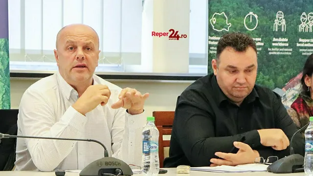 Un bărbat cu cămașă albă vorbește cu mâinile ridicate, în timp ce un alt bărbat cu cămașă neagră stă lângă el la o masă.