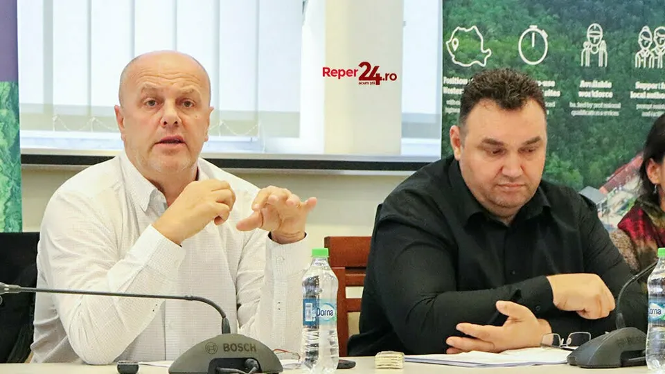 Un bărbat cu cămașă albă vorbește cu mâinile ridicate, în timp ce un alt bărbat cu cămașă neagră stă lângă el la o masă.