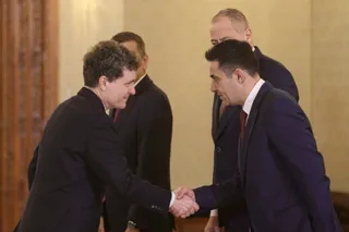 Patru bărbați îmbrăcați în costume închise stau într-o încăpere și doi dintre ei se strâng în mână.