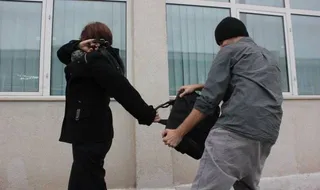 Un bărbat cu șapcă neagră și tricou gri ține o geantă neagră, în timp ce o persoană în haine negre stă cu spatele.