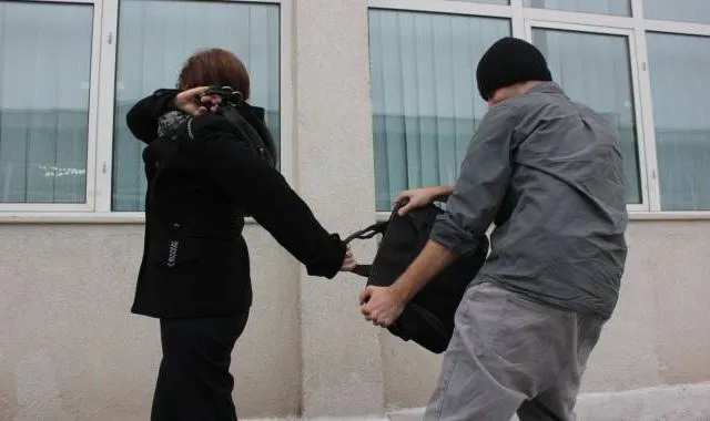 Un bărbat cu șapcă neagră și tricou gri ține o geantă neagră, în timp ce o persoană în haine negre stă cu spatele.