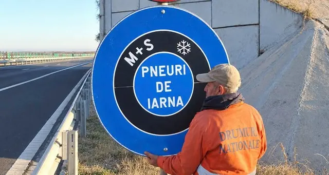 Un bărbat cu o căciulă și o vestă portocalie ține un semn circular albastru și negru pe marginea unei autostrăzi.