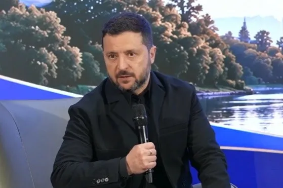 Un bărbat cu barbă și păr scurt, îmbrăcat în sacou negru, ține un microfon negru în mână în fața unui fundal cu copaci și apă.