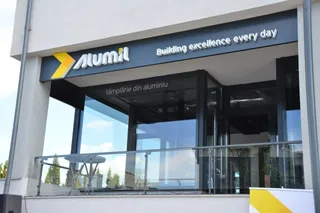 O clădire cu o suprafață vitrată mare prezintă un panou negru pe care este scris logo-ul Alumil și textul Building excellence every day.