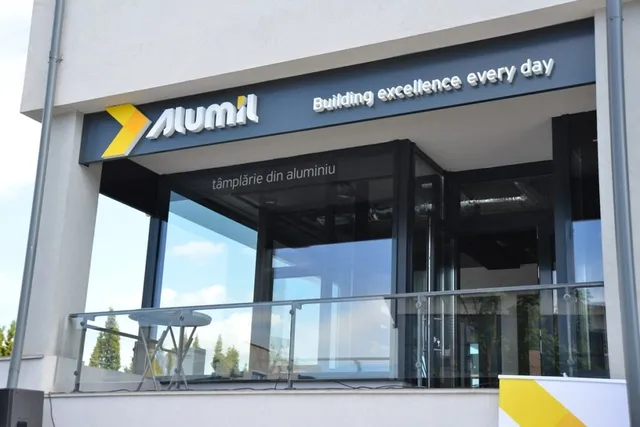 O clădire cu o suprafață vitrată mare prezintă un panou negru pe care este scris logo-ul Alumil și textul Building excellence every day.
