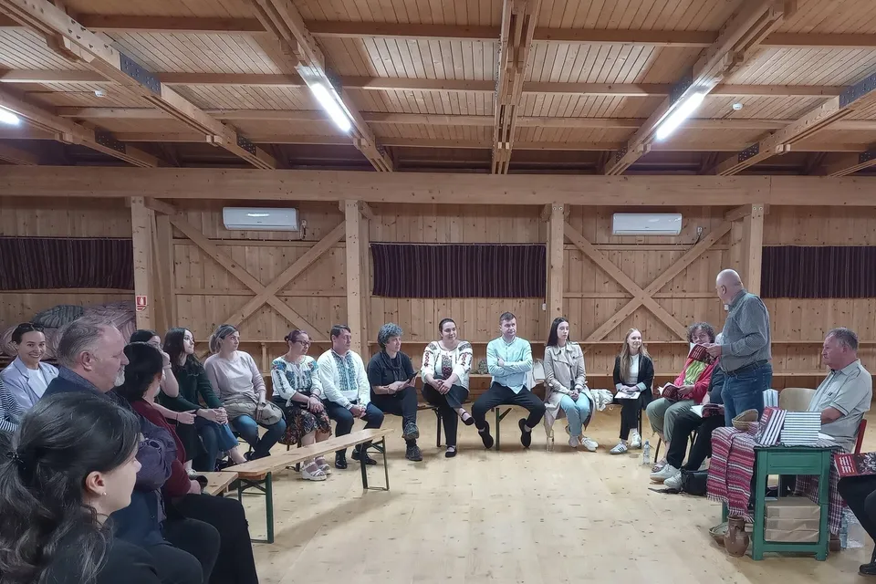 Un grup de persoane stă așezat pe scaune în semicerc într-o încăpere cu pereți și tavan din lemn, în timp ce un bărbat stă în picioare.