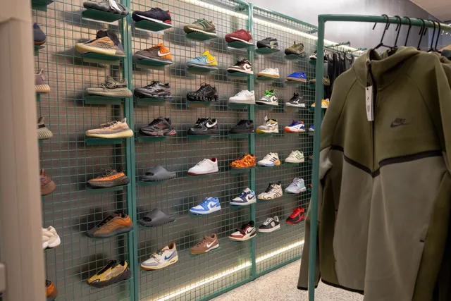 Pantofi sport de diverse culori și modele sunt expuși pe rafturi metalice verzi, lângă un hanorac de culoare verde măsliniu suspendat pe un umeraș.