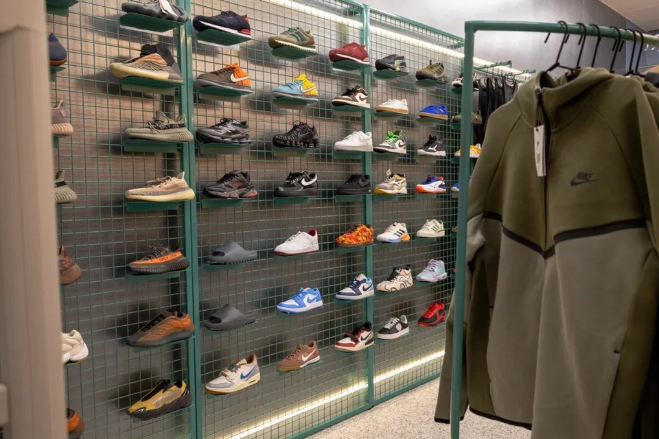 Pantofi sport de diverse culori și modele sunt expuși pe rafturi metalice verzi, lângă un hanorac de culoare verde măsliniu suspendat pe un umeraș.