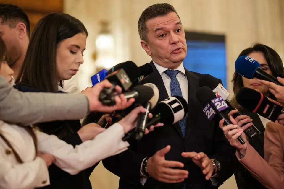 Un bărbat într-un costum închis și cravată albastră vorbește înconjurat de mai mulți microfoane ținute de persoane.