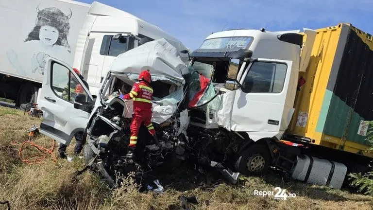 Un salvator în echipament roșu și galben se află lângă cabina unui camion alb zdrobit într-un accident rutier cu un alt vehicul mare.