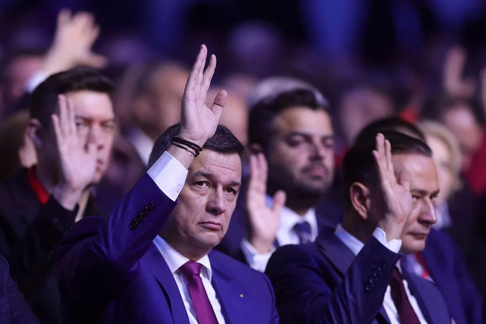 Mai mulți bărbați în costume și cravate ridică mâinile în semn de prezență într-o sală cu iluminare albastră.