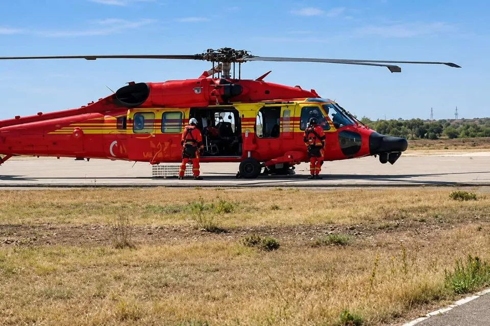 Un elicopter roșu cu dungi galbene este parcat pe o pistă de aterizare lângă un câmp de iarbă uscată sub un cer senin.