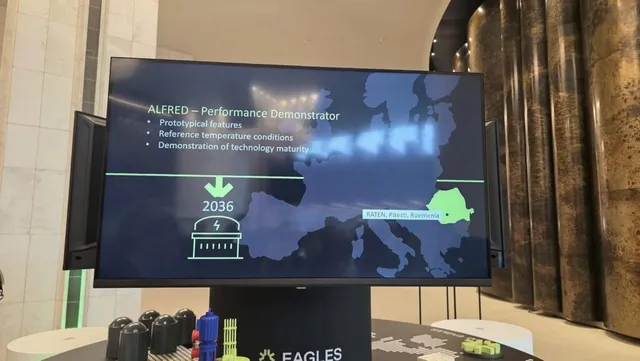 Un monitor afișează o hartă a Europei și text despre proiectul ALFRED, fiind poziționat pe un suport negru lângă niște obiecte colorate.