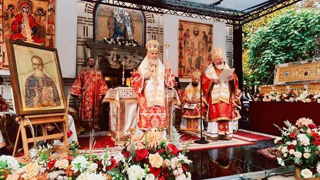 Imaginea arată un preot și un episcop în veștminte liturgice roșii și albe, stând în fața unui iconostas și a unui tablou religios, înconjurati de flori.