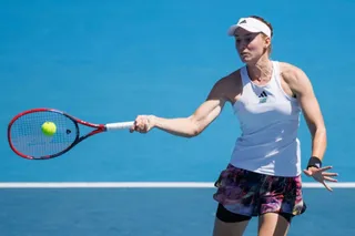 O femeie cu vizieră albă și tricou alb lovește o minge galbenă cu o rachetă de tenis pe un teren de culoare albastră sub un cer senin.